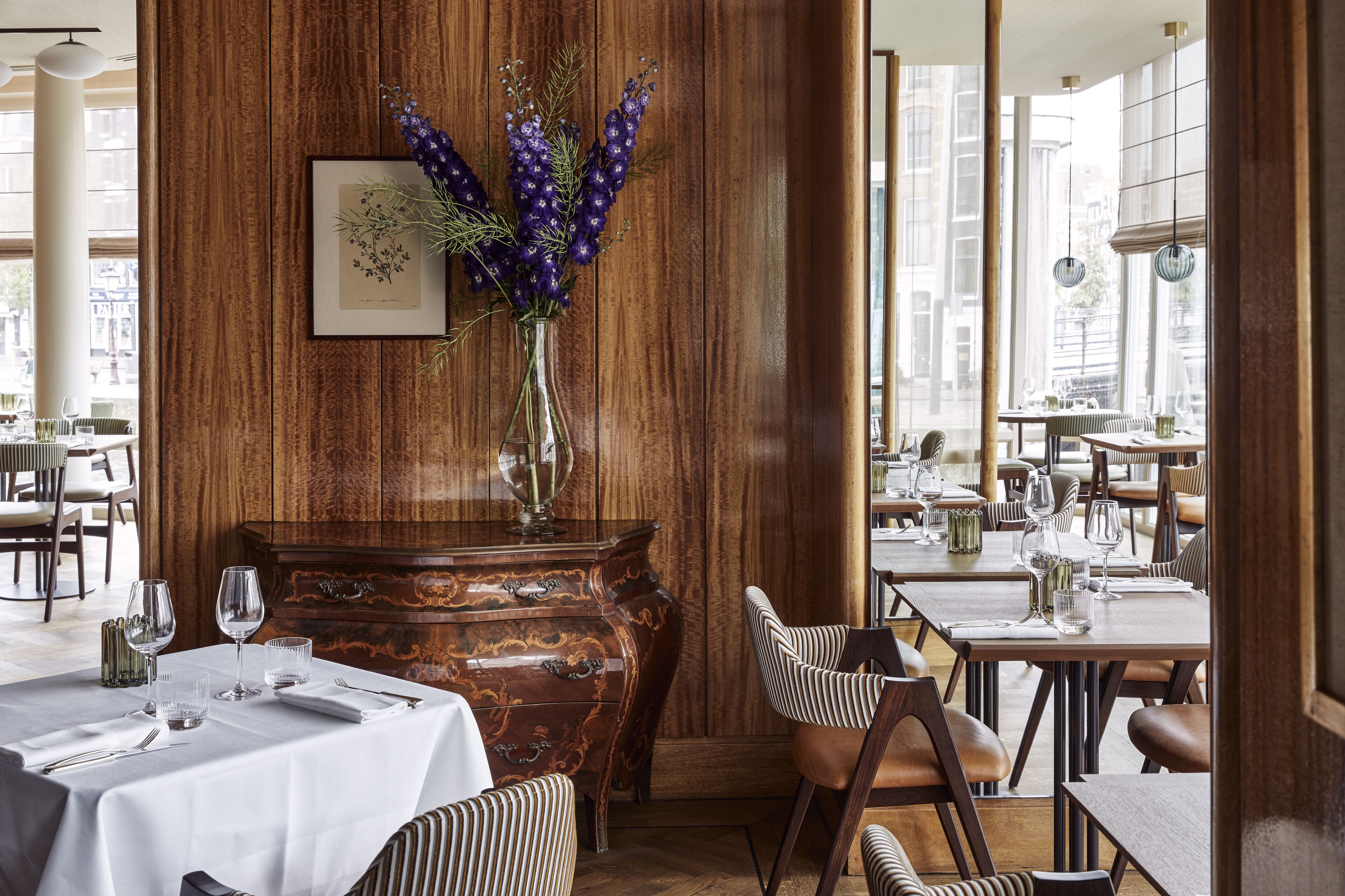 Un restaurant avec des murs lambrés, un vase rempli de fleurs violettes et des tables dressées pour le dîner.