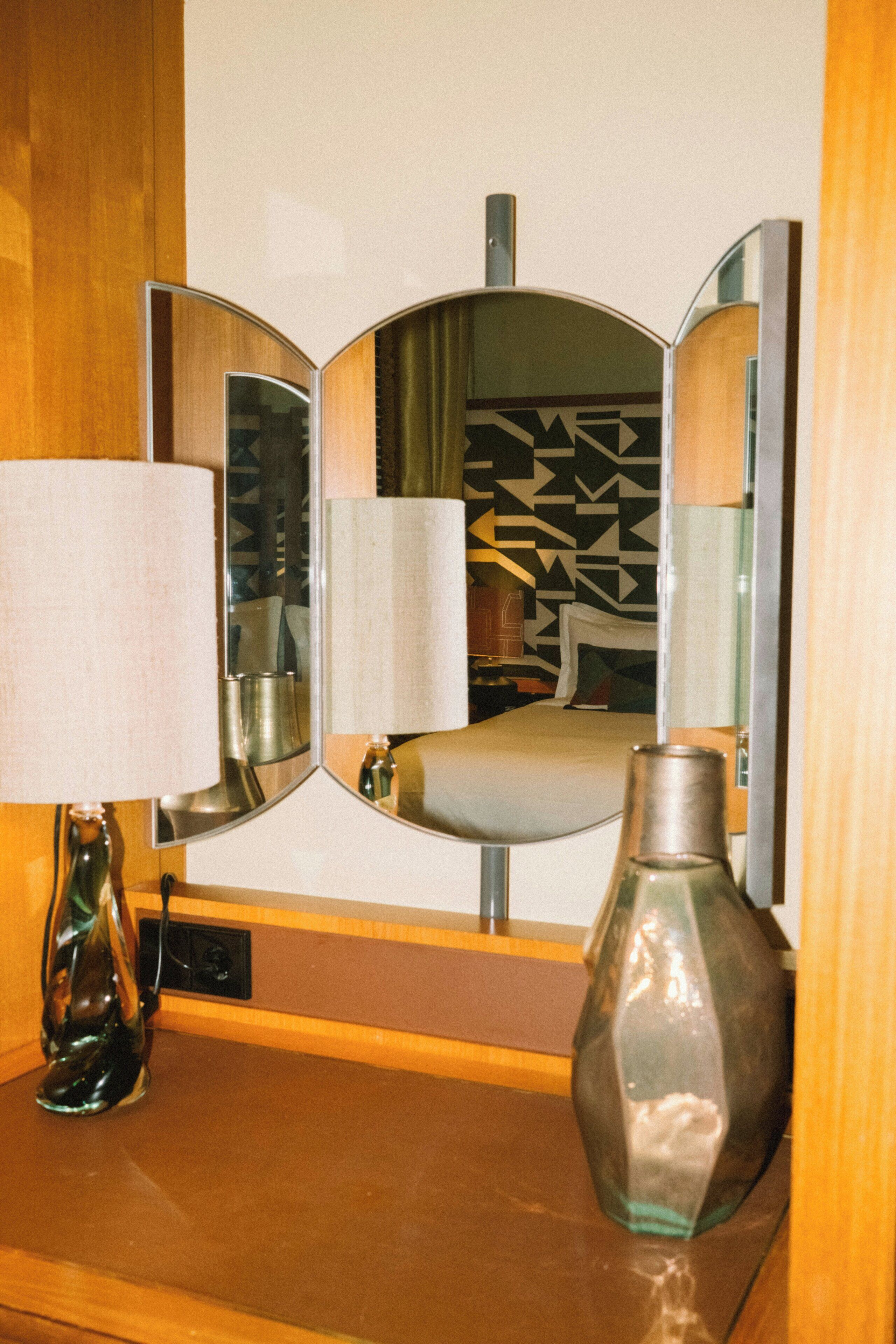 A mirror with a geometric pattern, a bedside table with a lamp, and a vase.