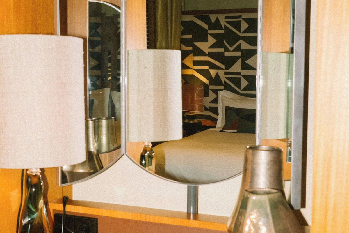 A mirror with a geometric pattern, a bedside table with a lamp, and a vase.