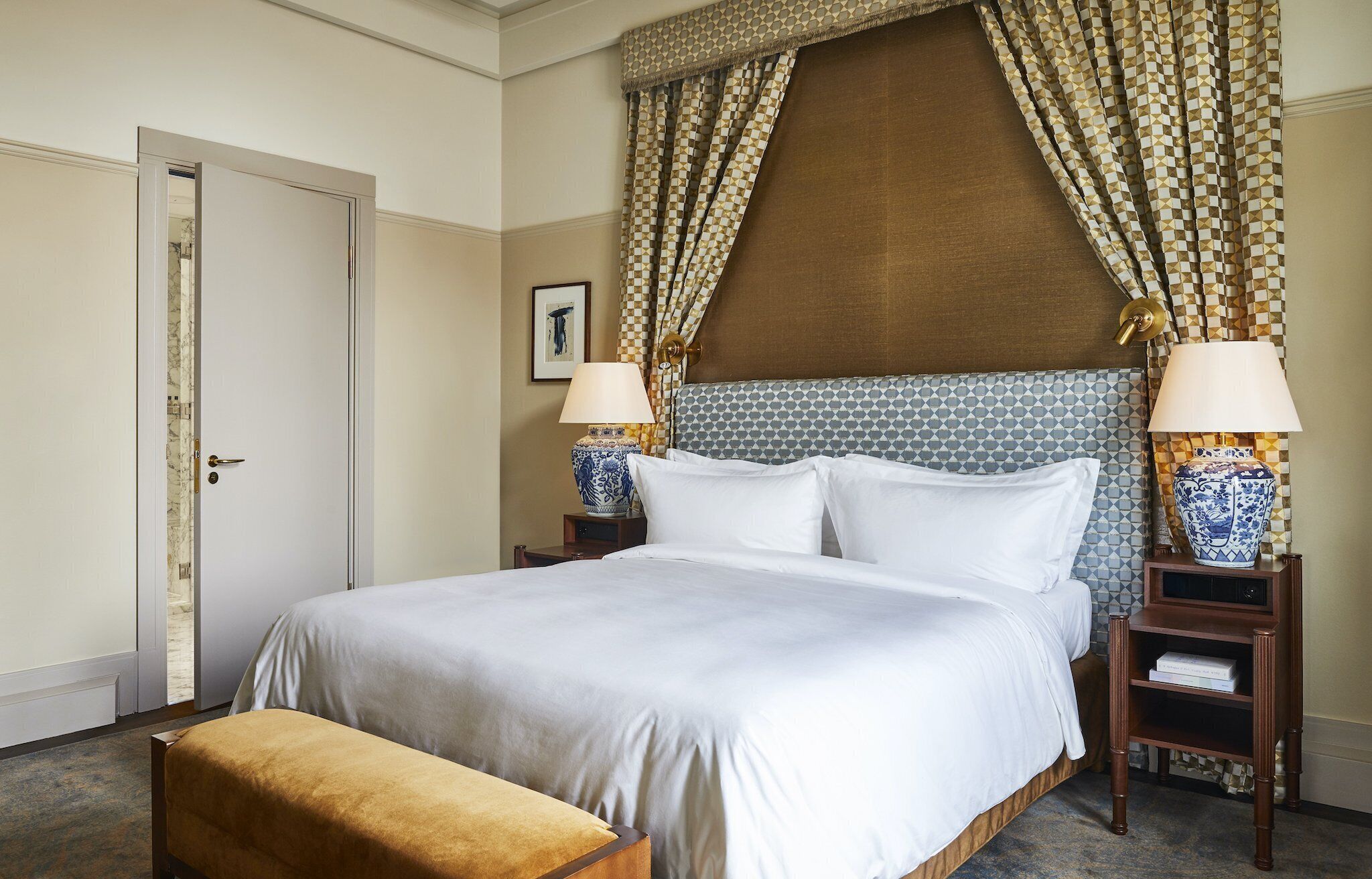 Een hotelkamer met een groot bed, twee nachtkastjes met lampen, een houten bankje en een deur.