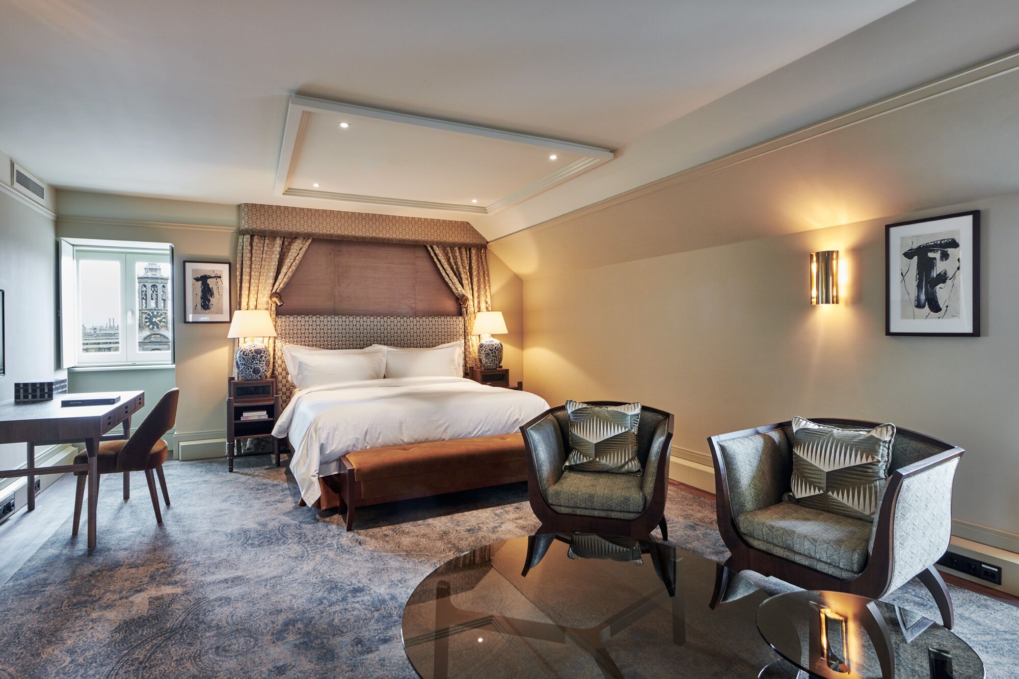 A hotel room with a large bed, two armchairs, a desk, and a view of a cityscape through the window.