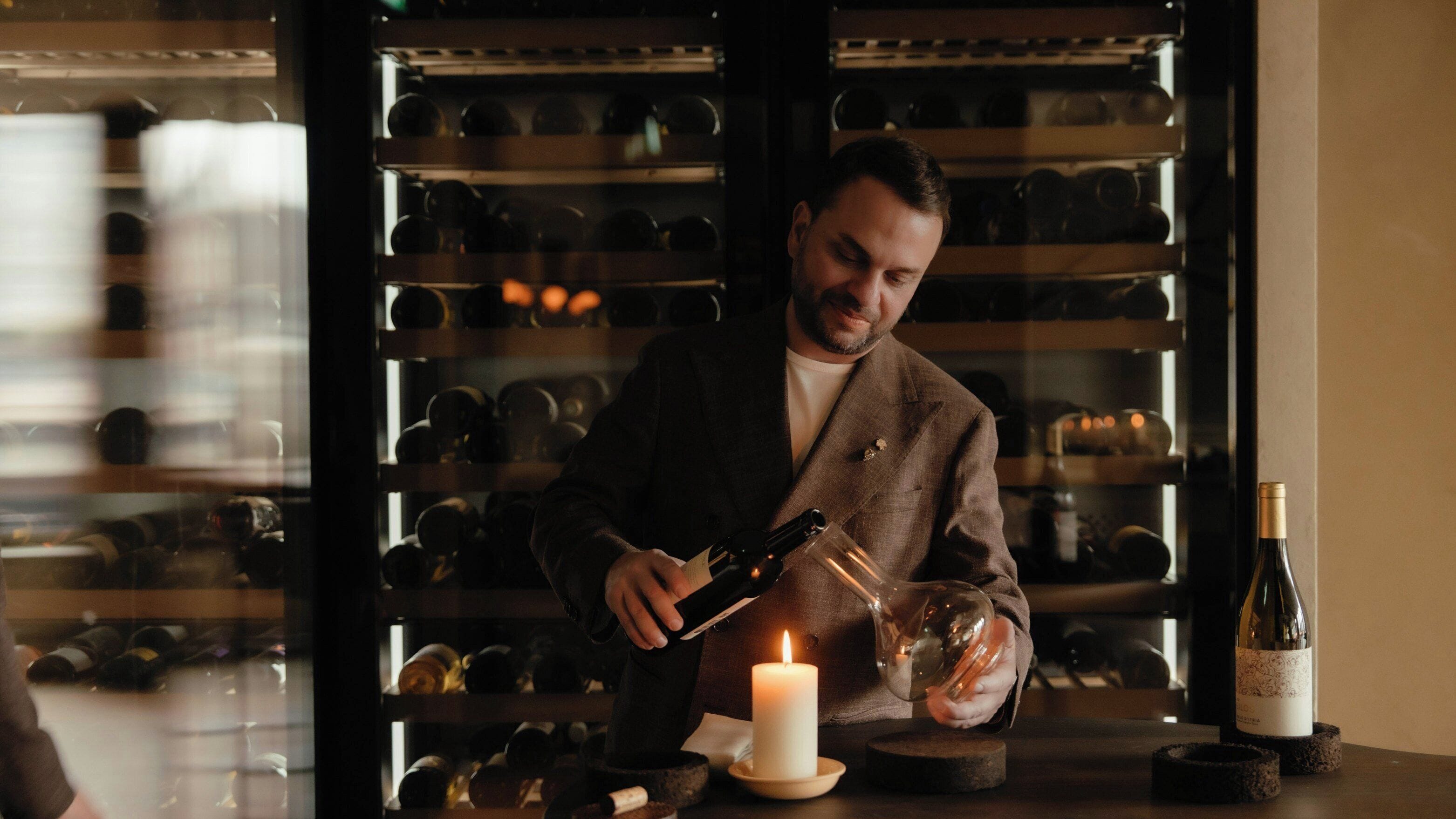 Una persona sirve vino de una botella en una copa, con una vela encendida sobre la mesa y botellas de vino al fondo.