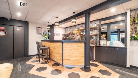 A bar area with a wooden counter, two wicker bar stools, hanging pendant lights, and a patterned wall with shelves displaying various items.. Kyriad Direct Limoges Nord