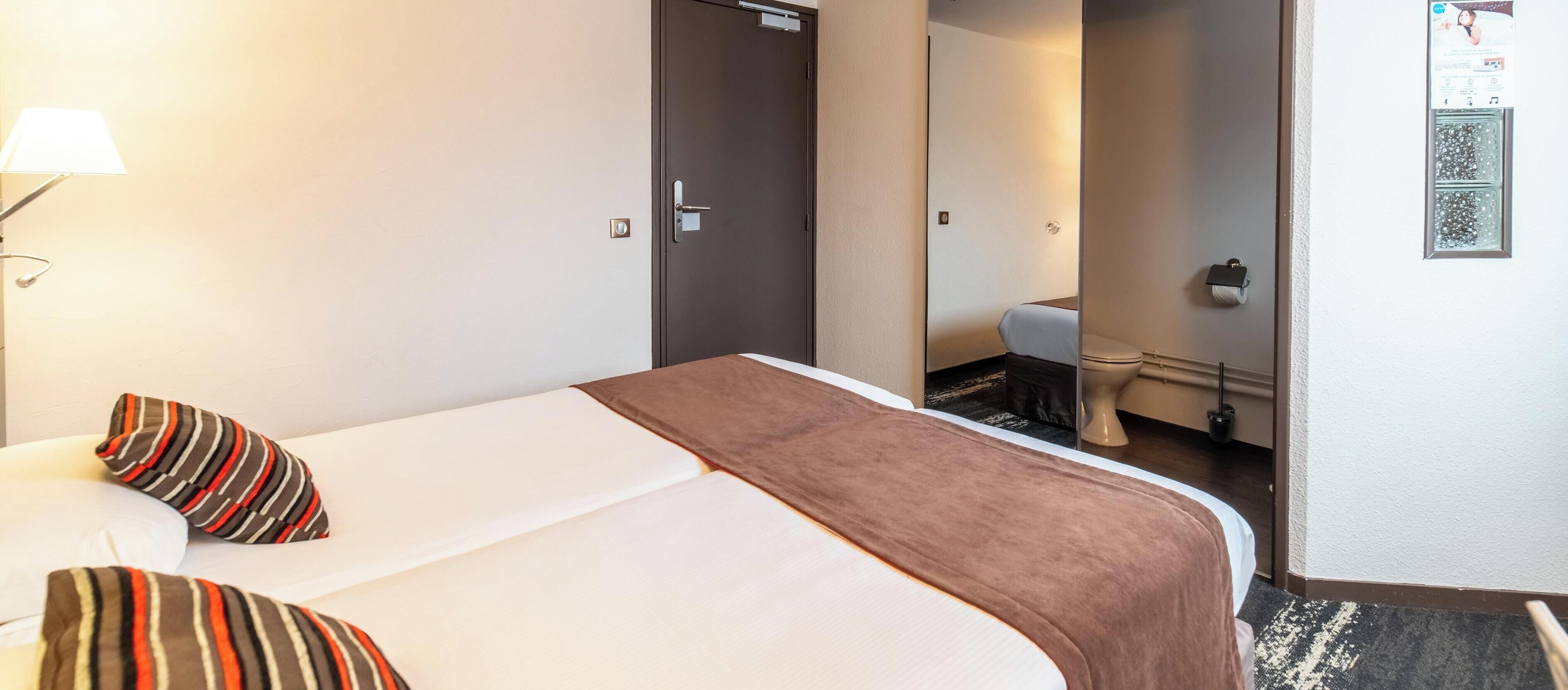 A hotel room with a bed, pillows, a lamp, a door, a mirror, and a toilet visible in the background.