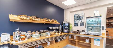 A breakfast bar with baskets of pastries, a coffee machine, and a refrigerator with beverages.