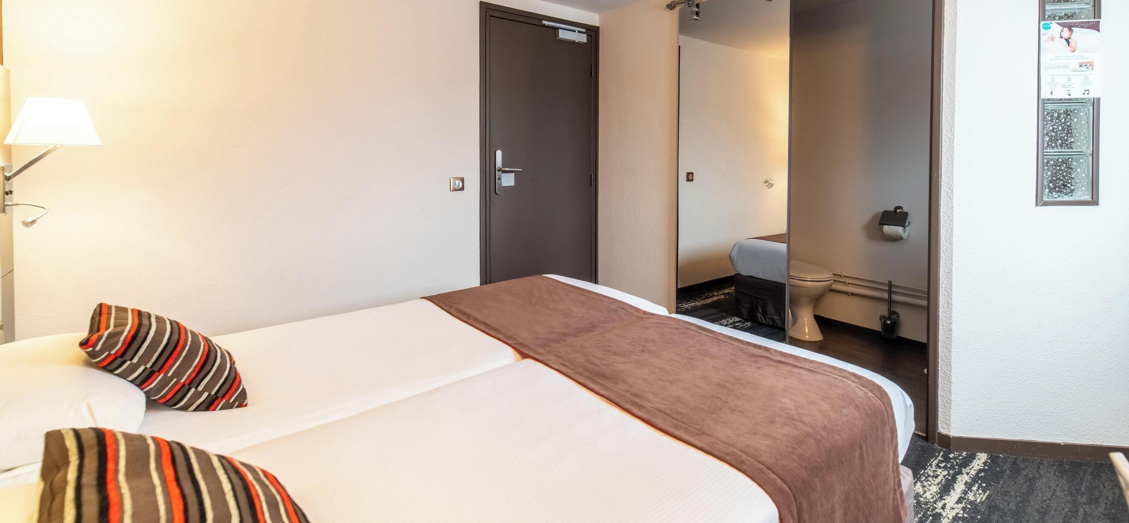A hotel room with a bed, a nightstand, a lamp, a mirror, and a bathroom visible through an open door.