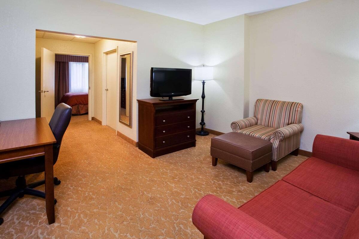 A hotel room with a wooden desk, a television on a stand, a striped armchair, a red sofa, and a lamp.