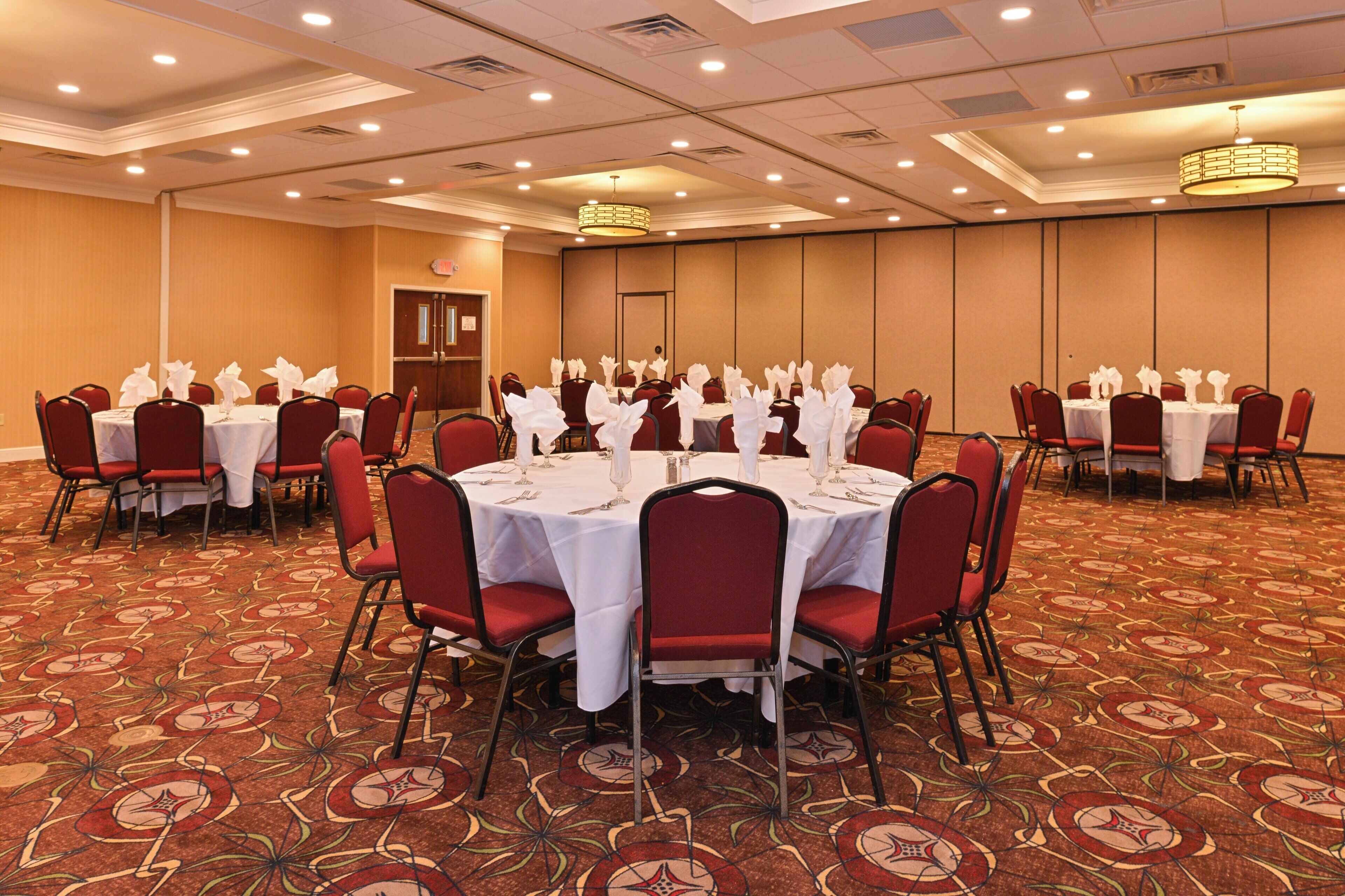 Une salle de banquet avec des tables rondes dressées pour le repas, des chaises rouges et un sol recouvert de moquette.