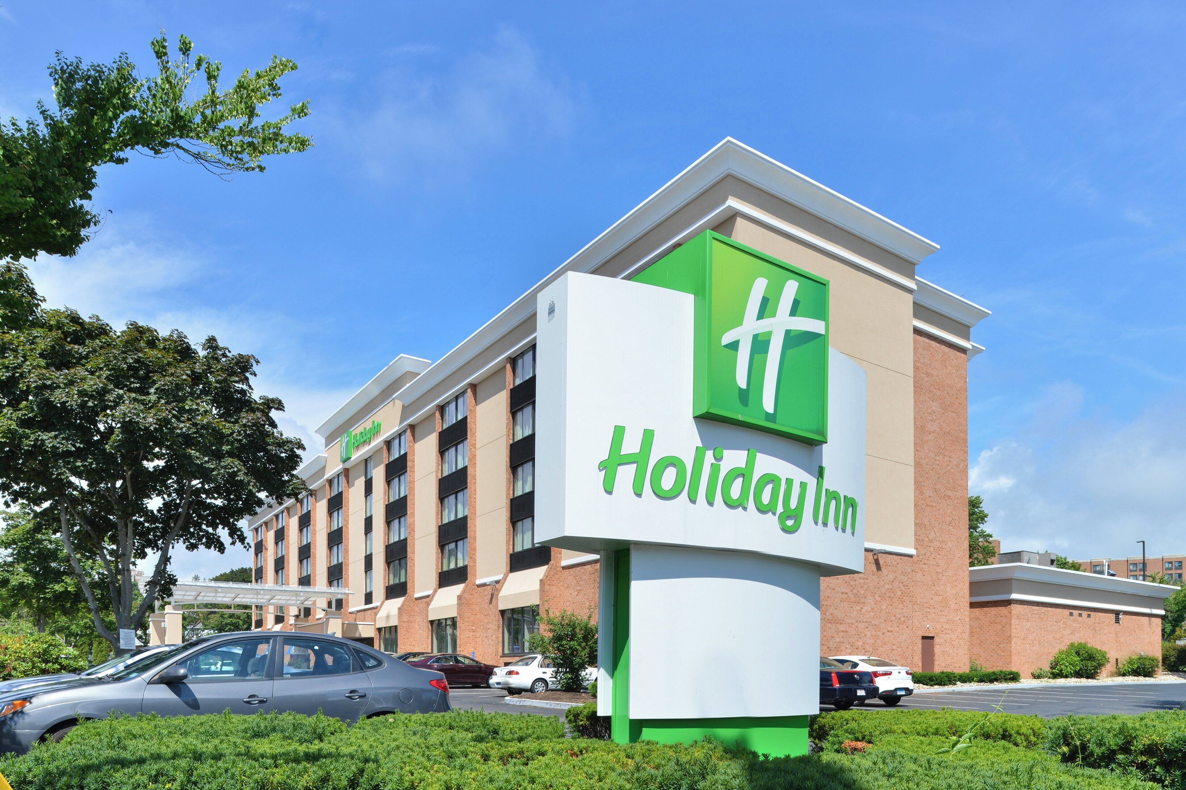 A Holiday Inn hotel with a prominent green and white sign, multiple floors, and a parking lot with cars.