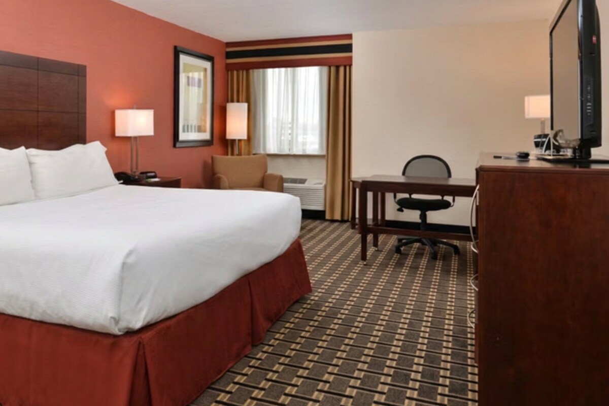 A hotel room with a large bed, a desk, a chair, a TV, and a window with curtains.