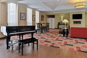 A hotel lobby with a grand piano, a red sofa, a patterned carpet, and a dining area with chairs and a table.