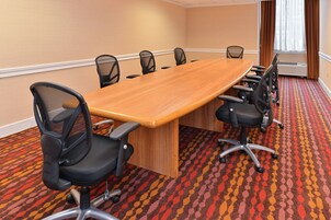 Un salón de conferencias con una larga mesa de madera rodeada por seis sillas de oficina negras con respaldo de malla. La sala tiene una alfombra estampada y paredes color beige.