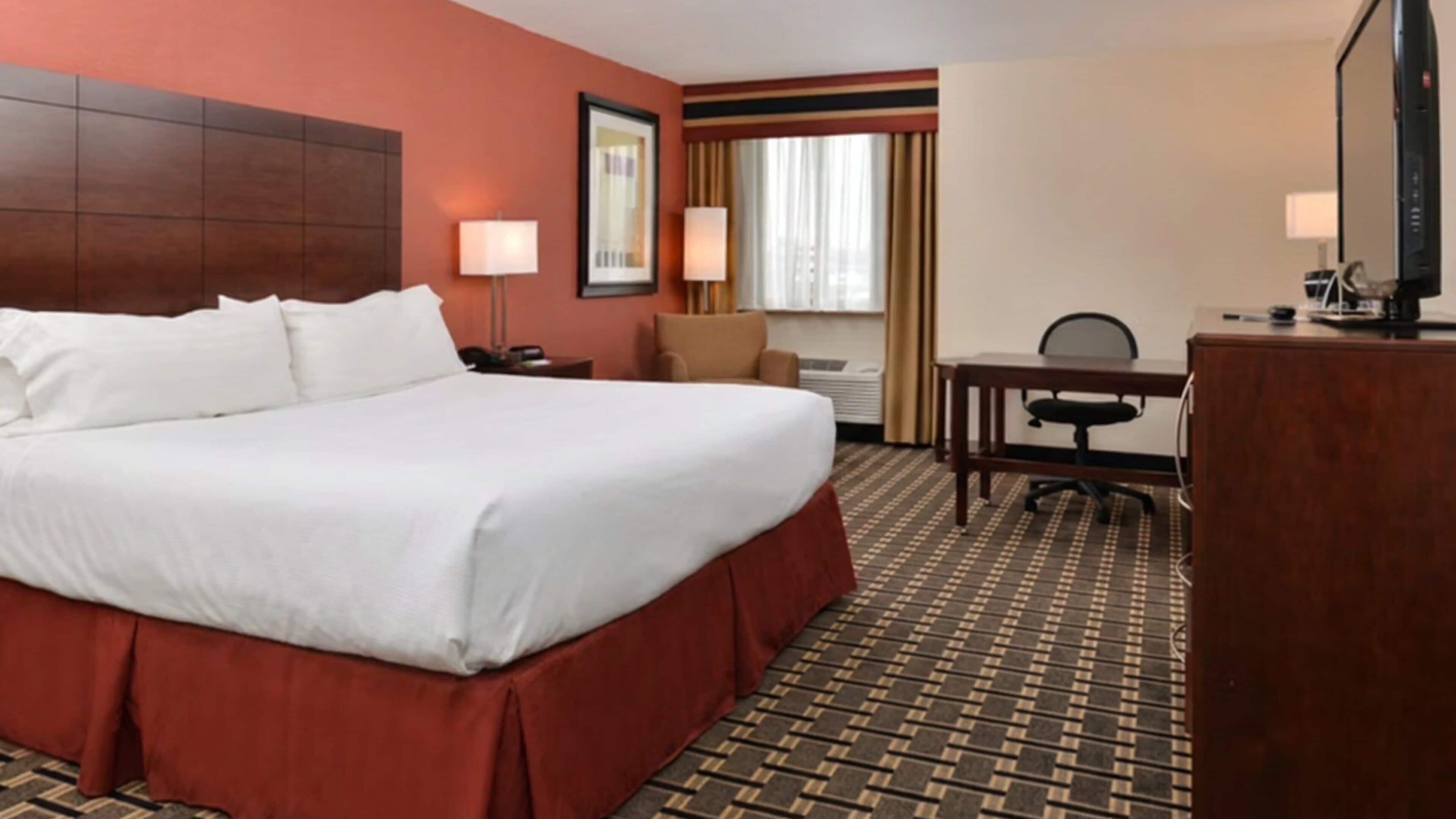 A hotel room with a large bed, a desk, a chair, a TV, and a window with curtains.
