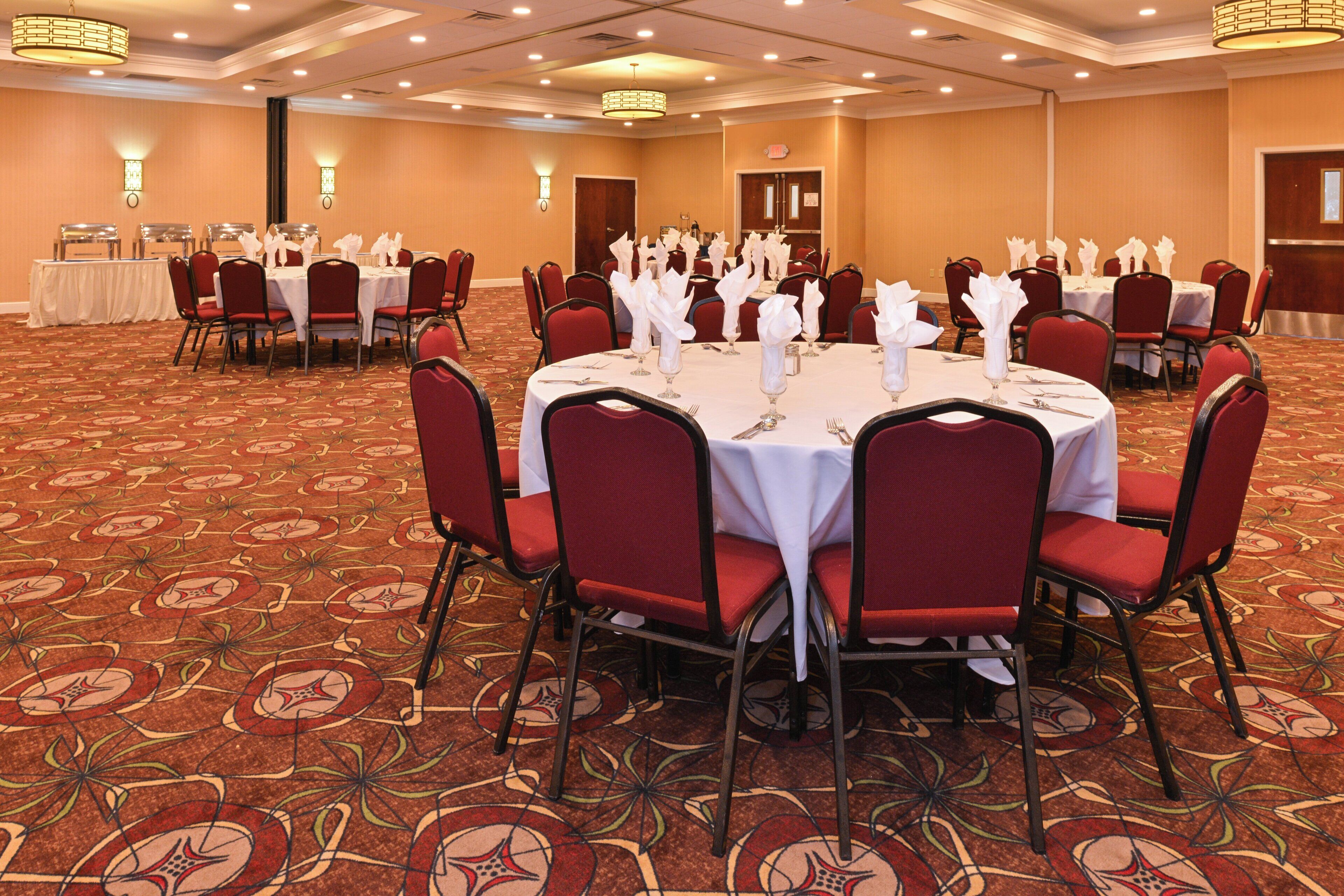 Un salón de banquetes con mesas redondas dispuestas para comer, sillas tapizadas en rojo y suelo alfombrado.