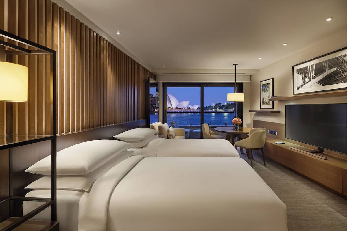 A hotel room with a large bed, a desk, a chair, a TV, and a view of the Sydney Opera House.