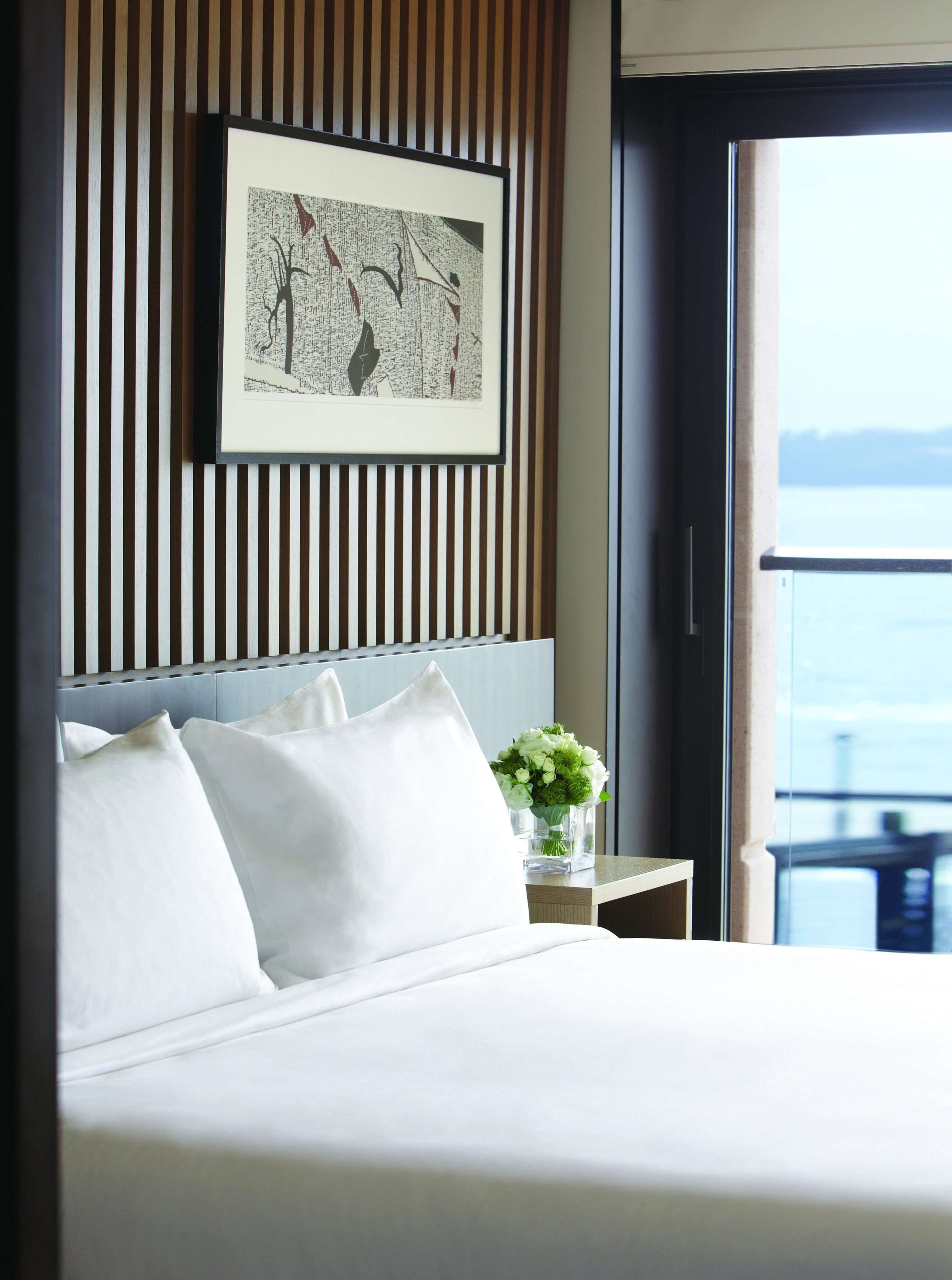 A hotel room with a bed, white pillows, a framed artwork on the wall, a nightstand with a vase of flowers, and a view of the sea through a window.