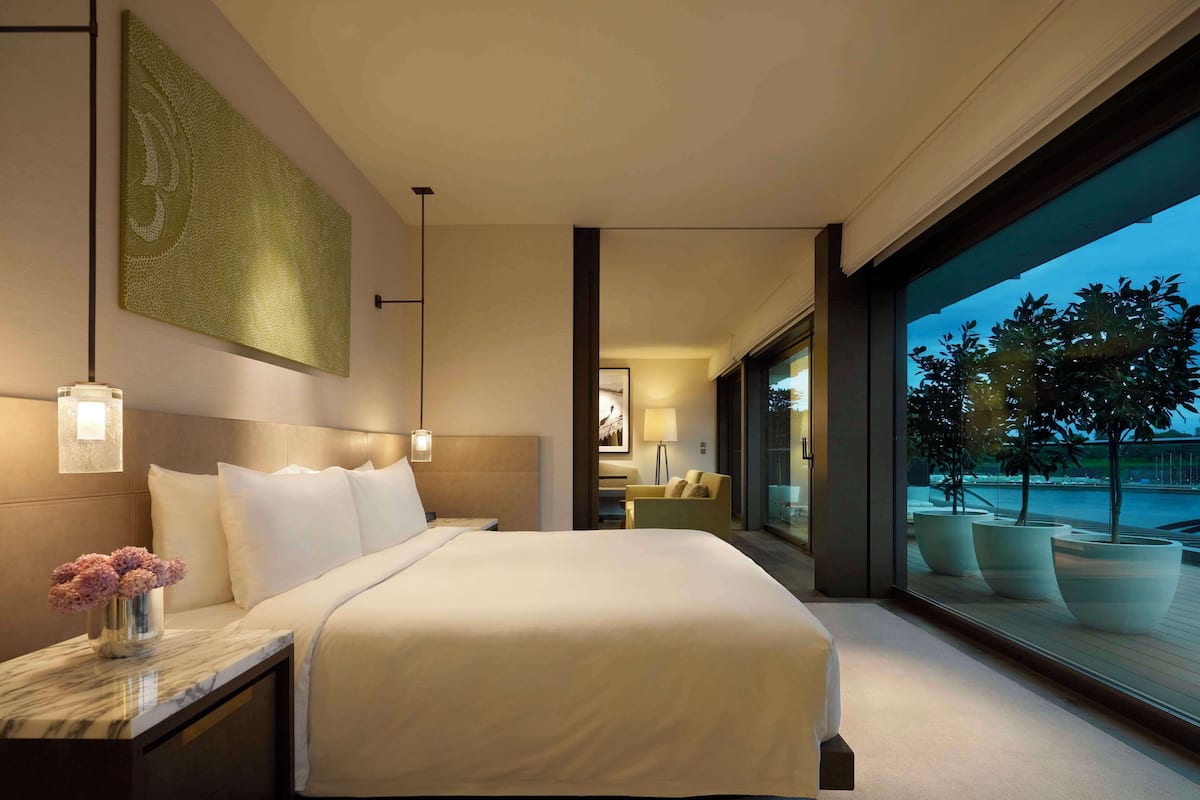 A modern hotel room with a large bed, a nightstand, a marble side table, a large window with a view of the outdoors, and a painting on the wall.