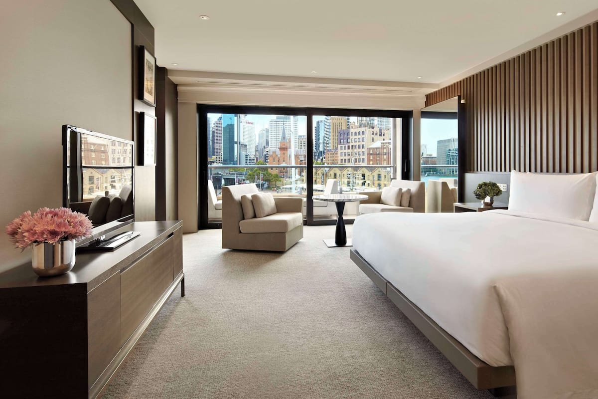 A modern hotel room with a large bed, a sofa, a desk, and a view of the cityscape through large windows.