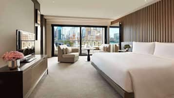 A modern hotel room with a large bed, a sofa, a desk, and a view of the cityscape through large windows.