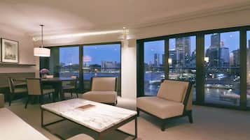 A modern hotel room with a large city view, a dining area with a table and chairs, and a seating area with two armchairs.