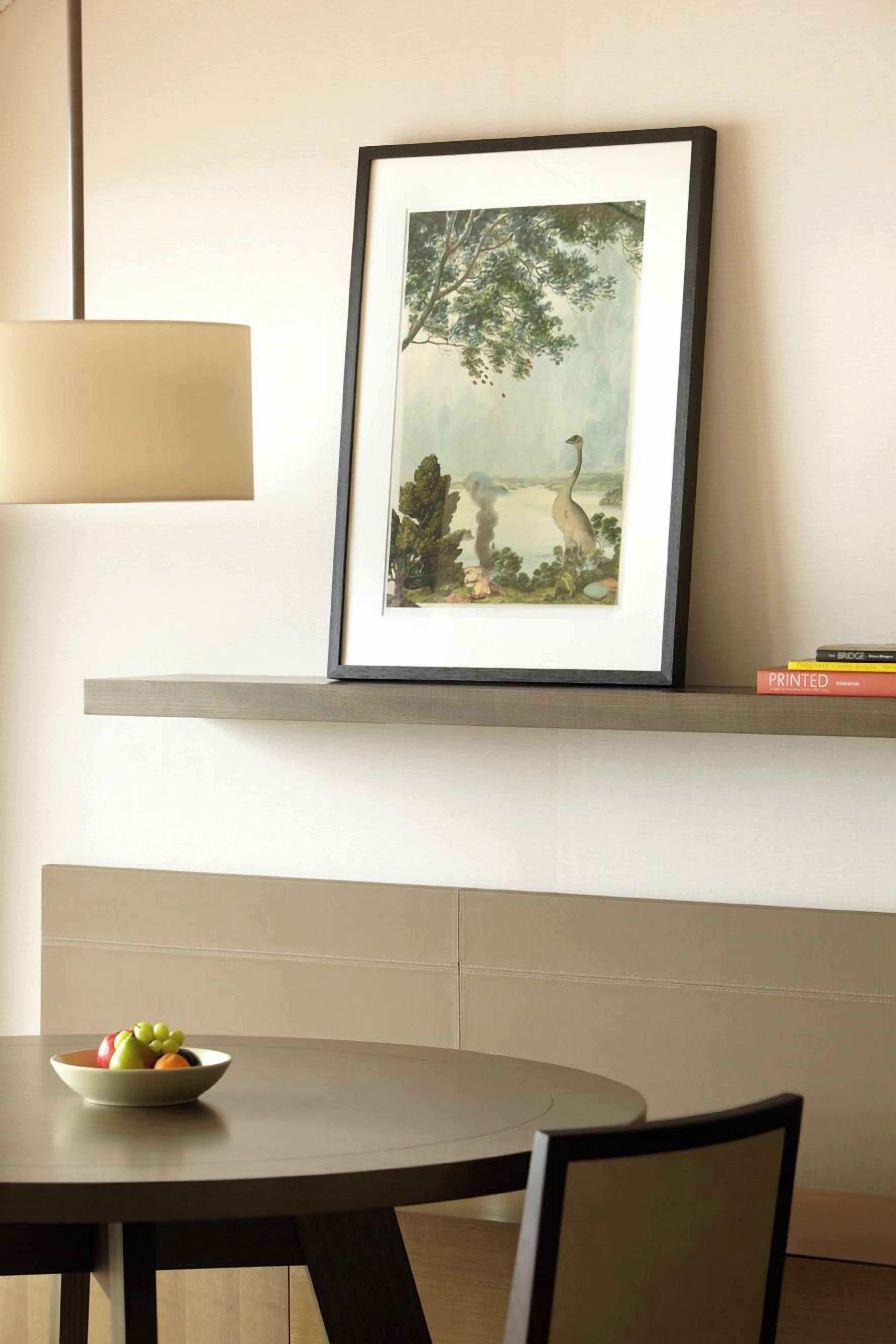 A framed artwork of a tree and bird on a wall, a bowl of fruit on a table, and a stack of books on a shelf.