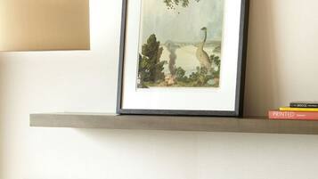 A framed artwork of a tree and bird on a wall, a bowl of fruit on a table, and a stack of books on a shelf.