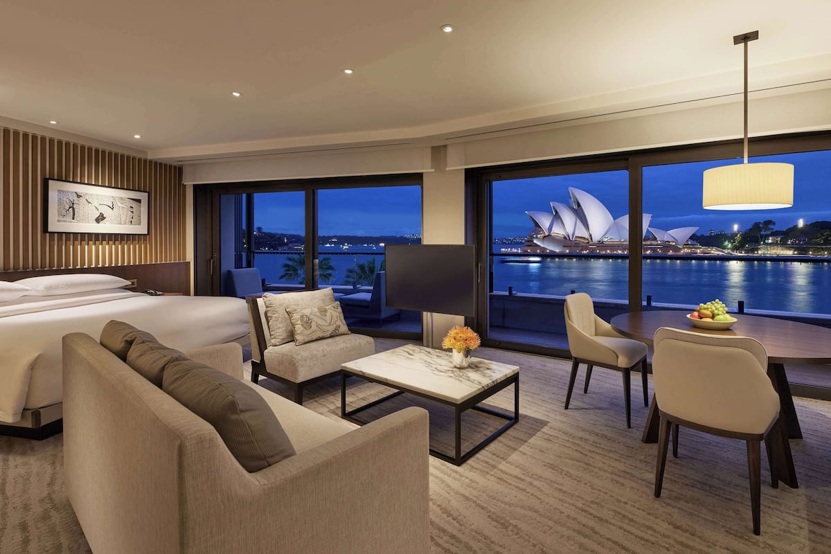 A modern hotel room with a large bed, a sofa, a coffee table with flowers, and a view of the Sydney Opera House.