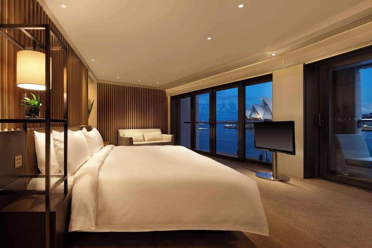 A modern hotel room with a large bed, a sofa, a desk with a computer, and a large window offering a view of a building and the sea.