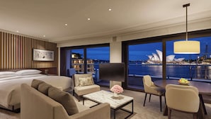 Habitación de hotel moderna con una cama grande, un sofá, una mesa de centro con un florero y un área de comedor con sillas. Un ventanal ofrece vistas de la Ópera de Sydney.