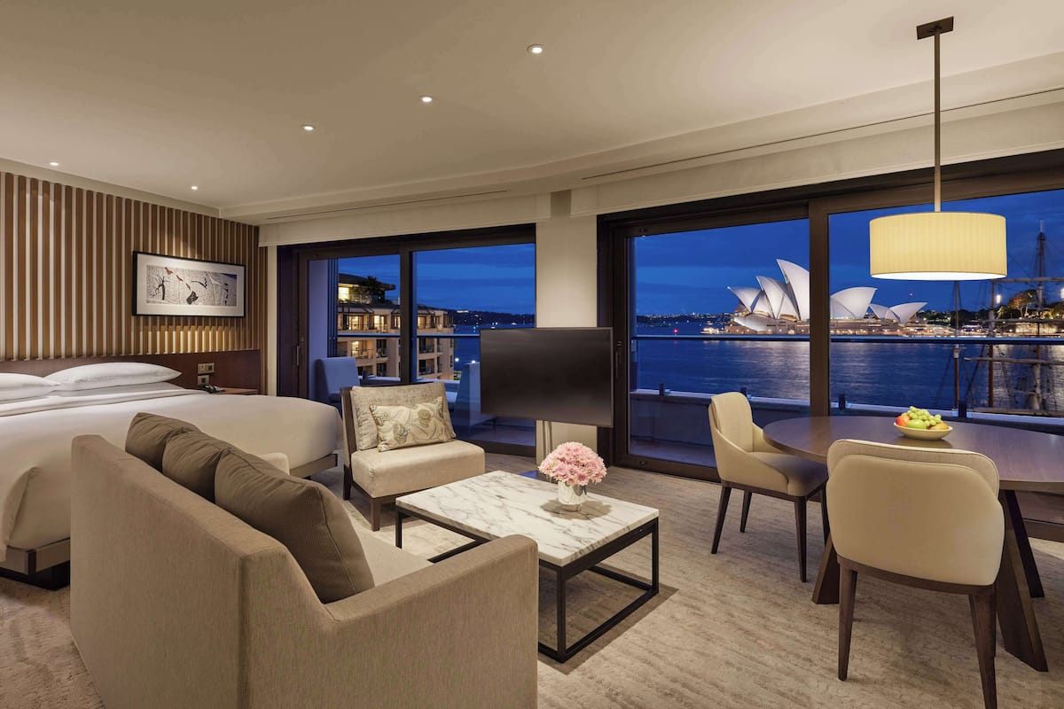 A modern hotel room with a large bed, a sofa, a coffee table with a vase, and a dining area with chairs. A large window offers a view of the Sydney Opera House.