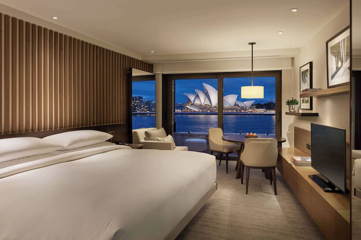 A hotel room with a large bed, a desk with a TV, a chair, and a view of the Sydney Opera House.