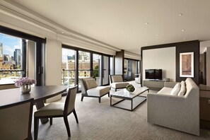 A modern living room with a sofa, chairs, a coffee table, and a TV. Large windows offer a city view.