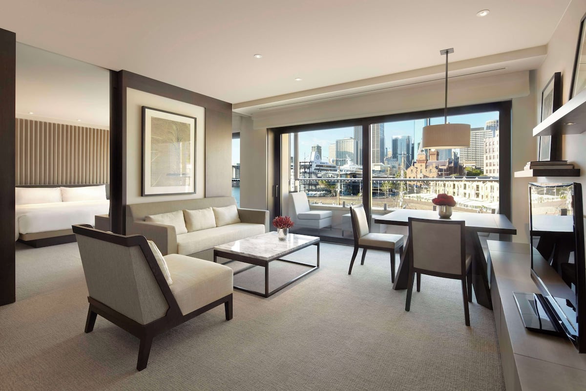 A modern hotel room with a large window offering a city view, a sofa, a coffee table, and a desk with a chair.