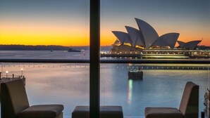Vista desde una habitación con un ventanal grande que da a un cuerpo de agua y la Ópera de Sídney al atardecer.