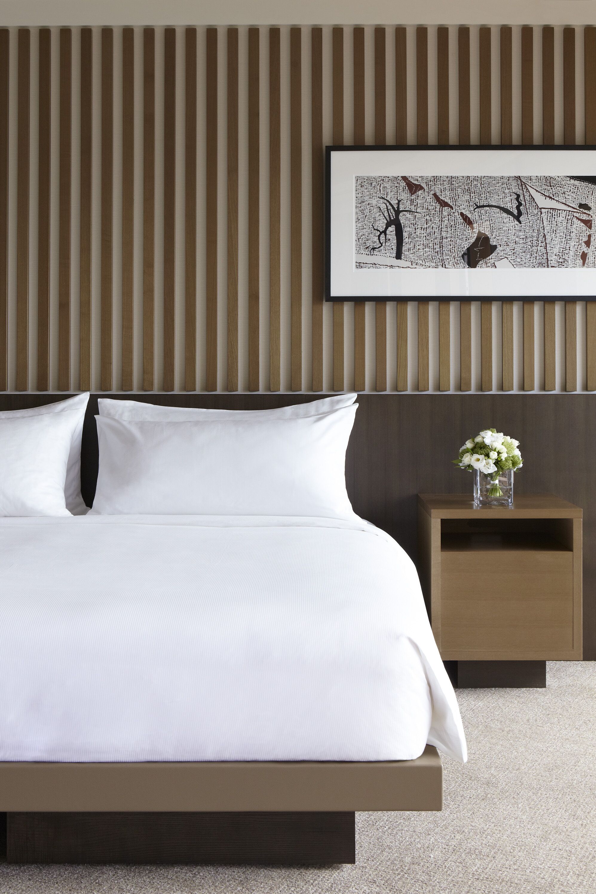 Una cama bien hecha con sábanas blancas, cabecera de madera, mesita de noche con un florero y un cuadro en la pared.
