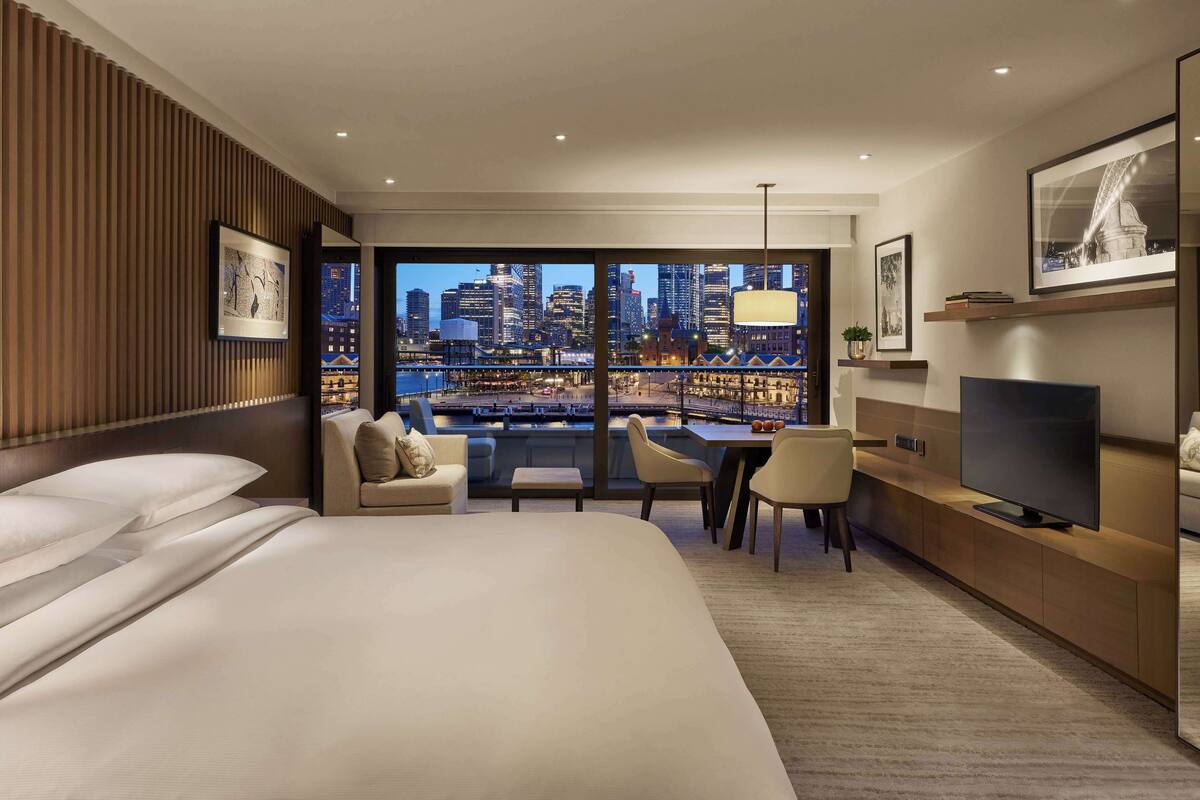 A modern hotel room with a large bed, a sofa, a desk, and a television. The room features a large window with a city view.