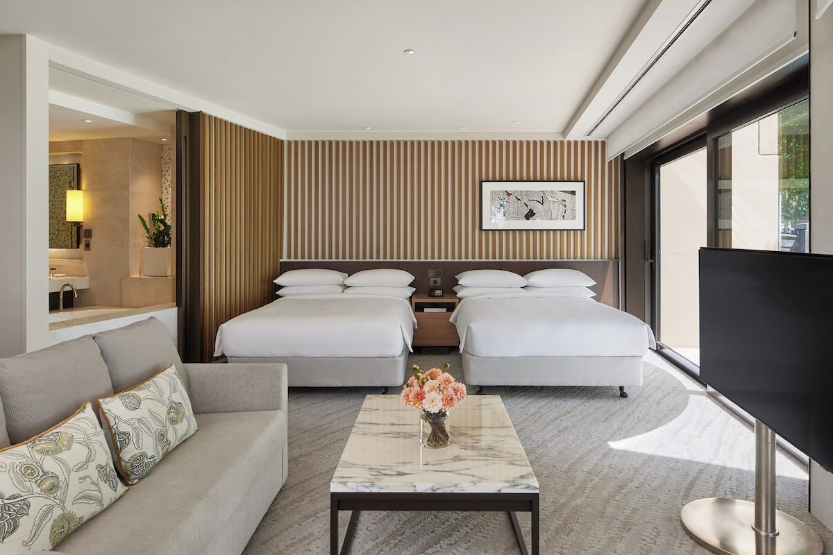 A hotel room with two beds, a sofa, a coffee table with flowers, a TV, and a large window.