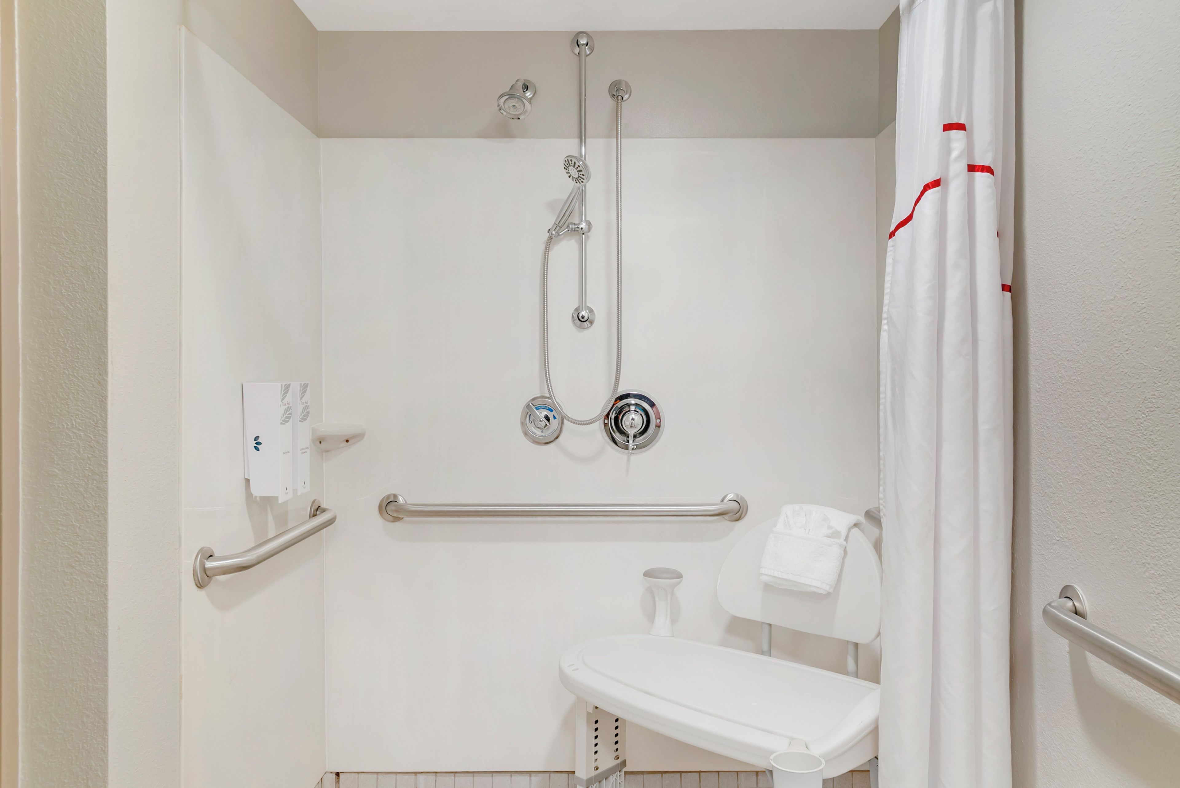 Un baño con ducha, barras de apoyo, secamanos y un inodoro.