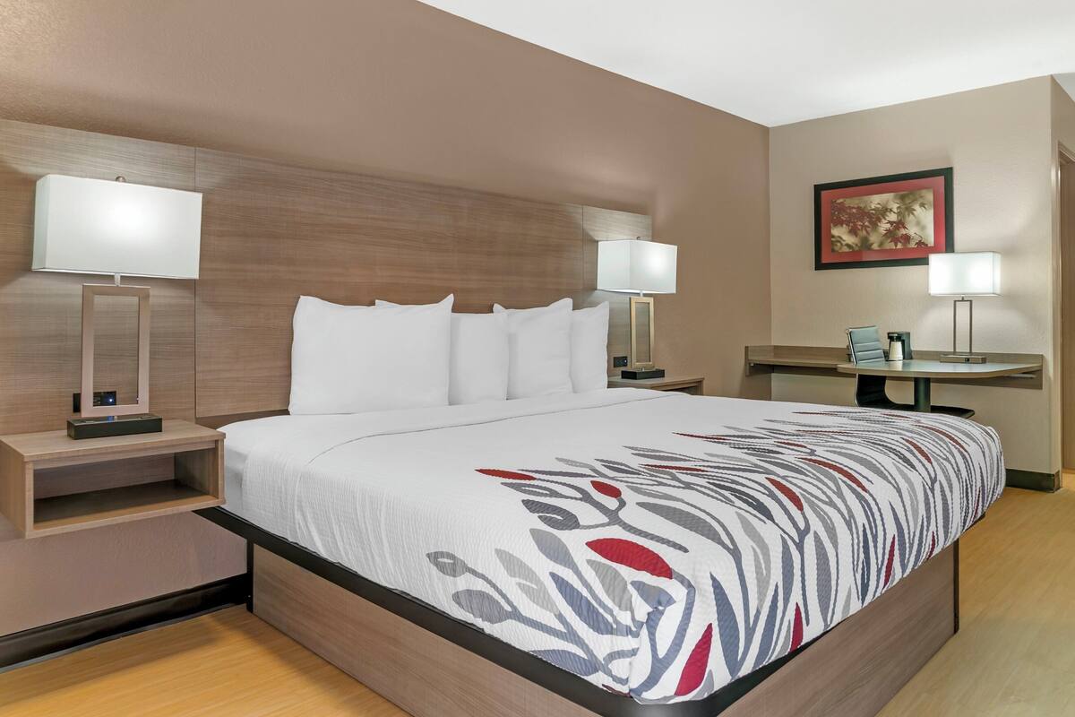 A modern hotel room with a large bed, two bedside tables with lamps, a small desk with a chair, and a framed picture on the wall.