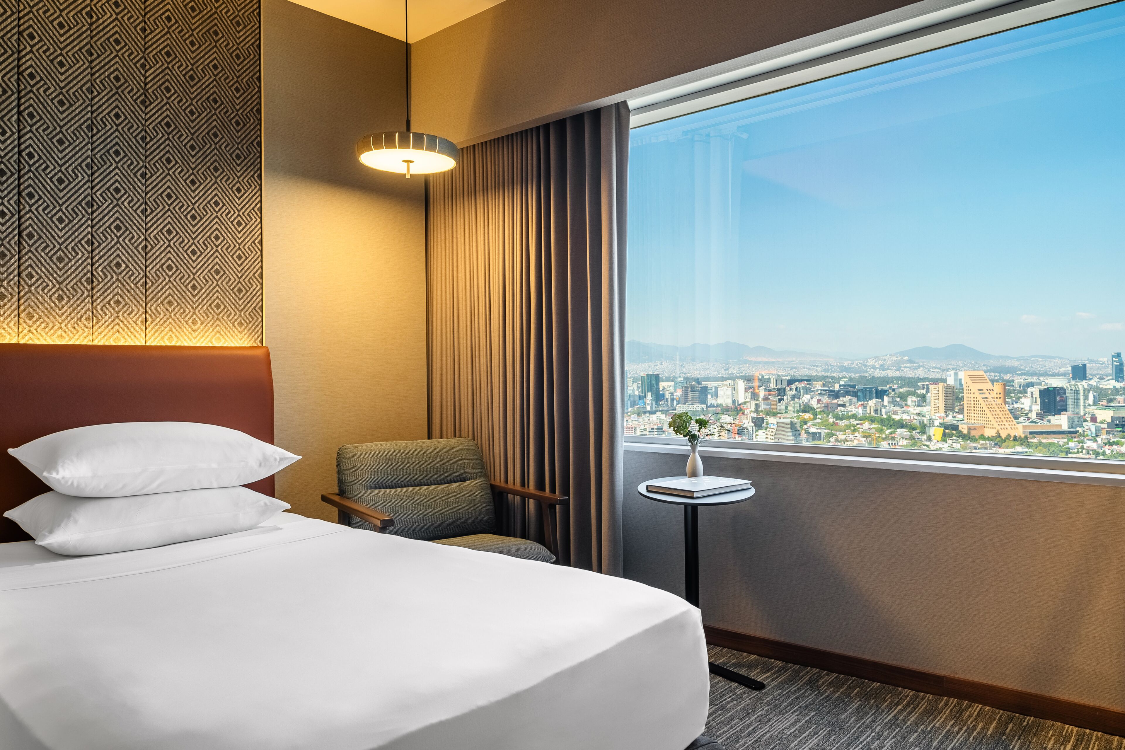Een hotelkamer met een groot bed, een stoel, een klein tafeltje en uitzicht op een stadsgezicht door een groot raam.