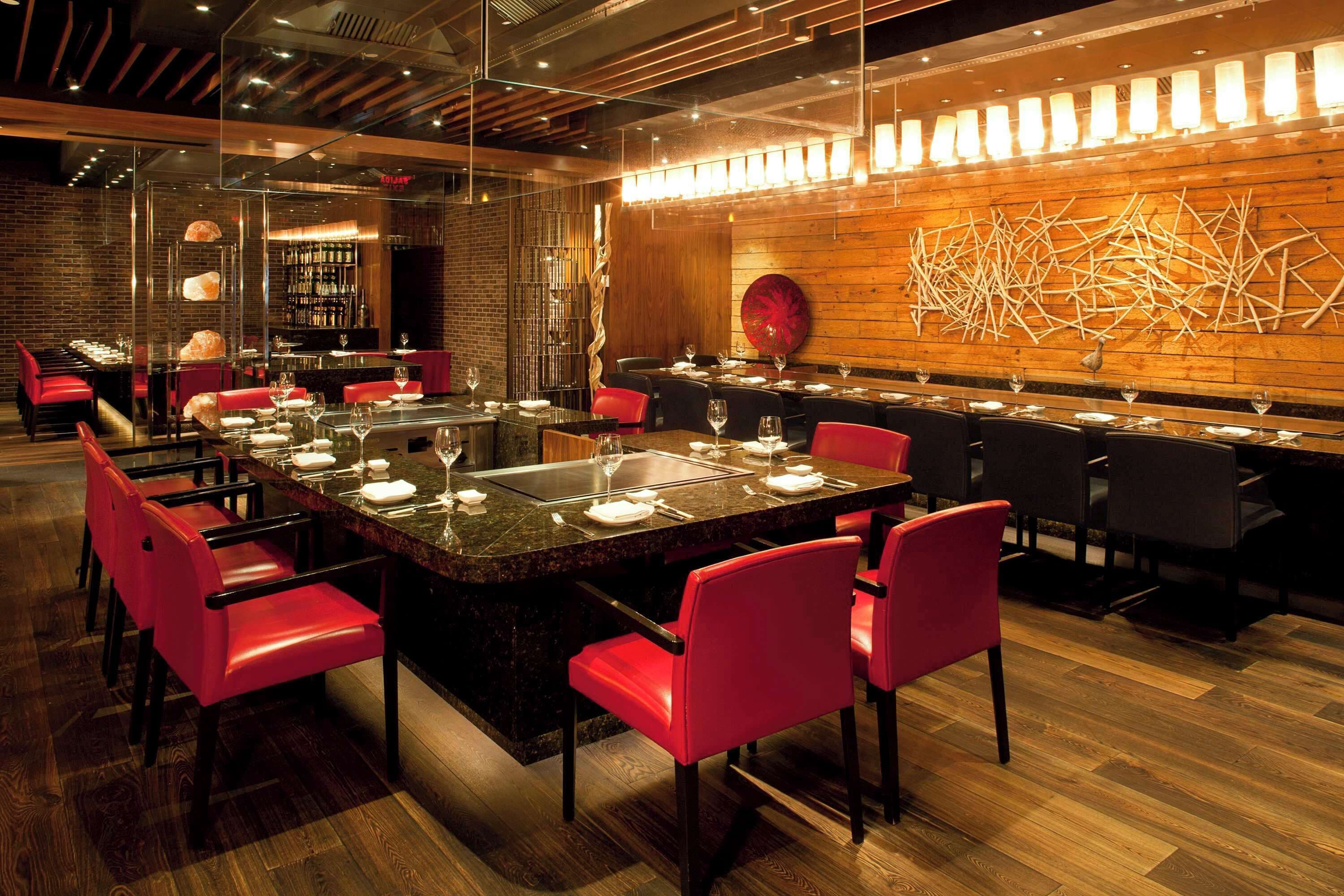 Un restaurante moderno con una mesa central rodeada de sillas rojas, una zona de bar con estantes llenos de botellas y una pared de madera con arte abstracto.