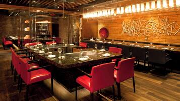 Un restaurante moderno con una mesa central rodeada de sillas rojas, una zona de bar con estantes llenos de botellas y una pared de madera con arte abstracto.