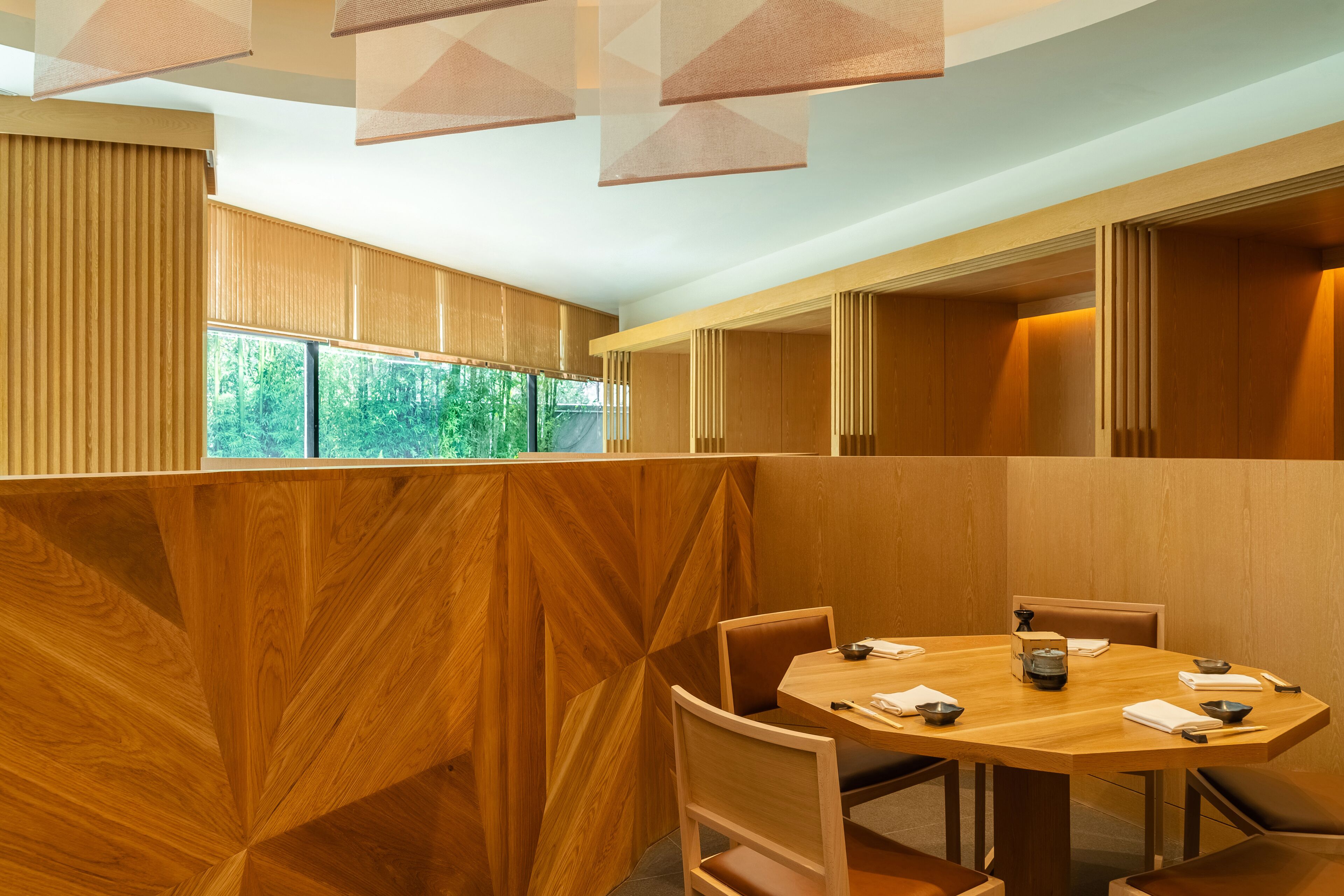 A modern dining area with a round wooden table, cushioned chairs, and a view of greenery through large windows.