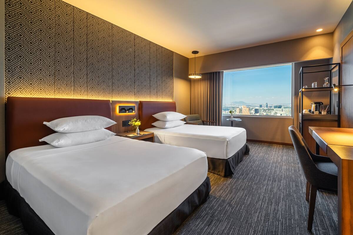 Une chambre d’hôtel avec deux lits, un bureau, une chaise, une grande fenêtre donnant sur la ville et un mur décoré de motifs.