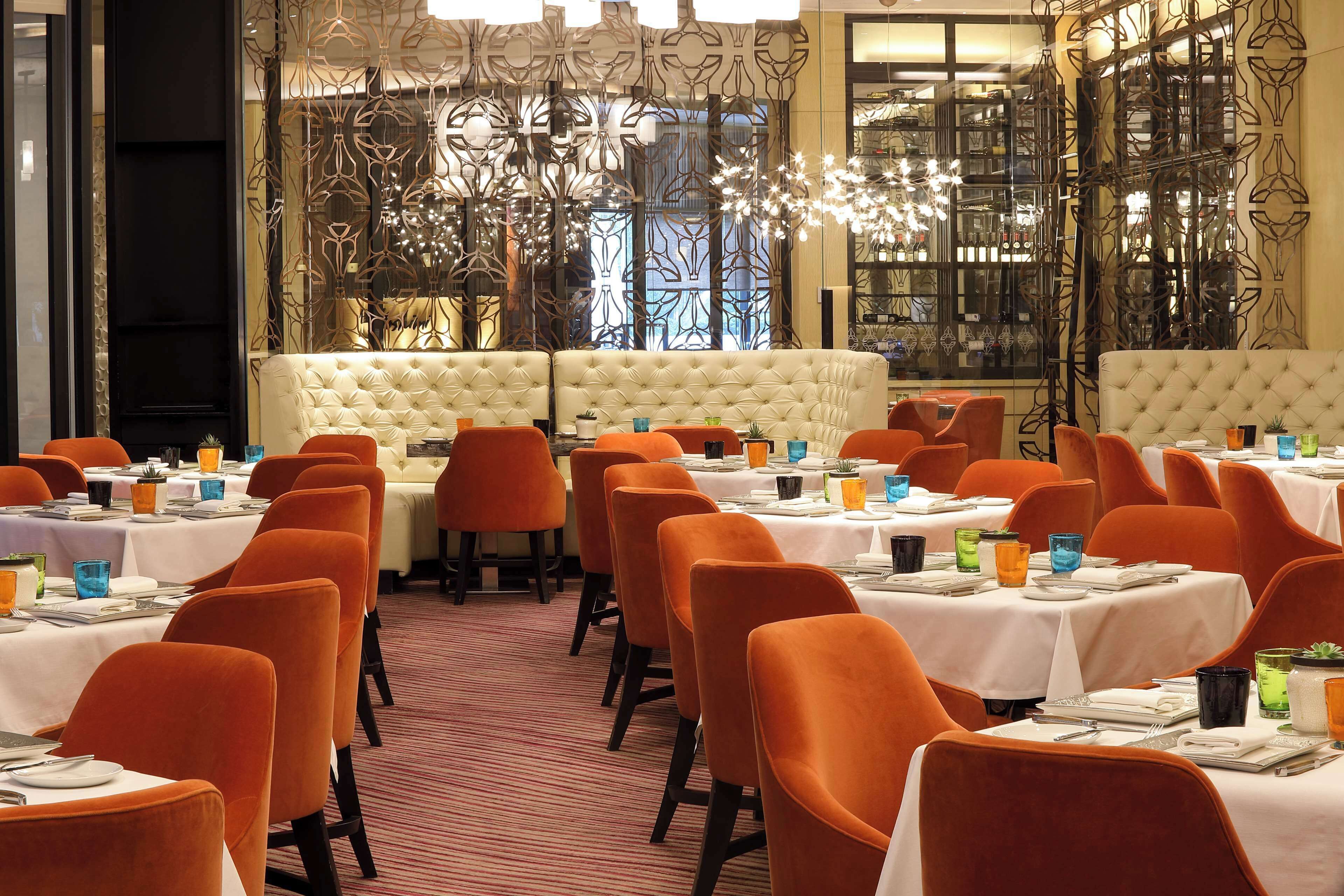 Un restaurante con sillas anaranjadas y mesas blancas, un sofá capitonado y detalles de metal decorativo.