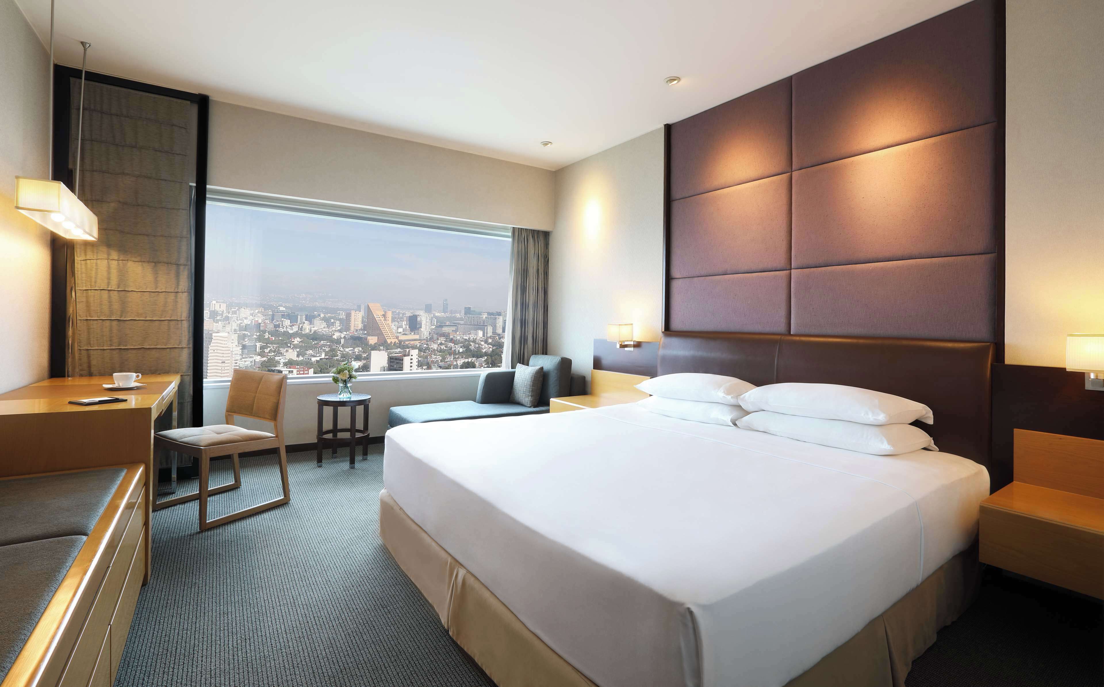 A hotel room with a large bed, a desk, a chair, a small table, and a view of the city.