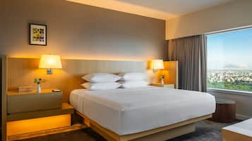 A hotel room with a large bed, two bedside tables, a nightstand, a view of the city, and a framed picture on the wall.