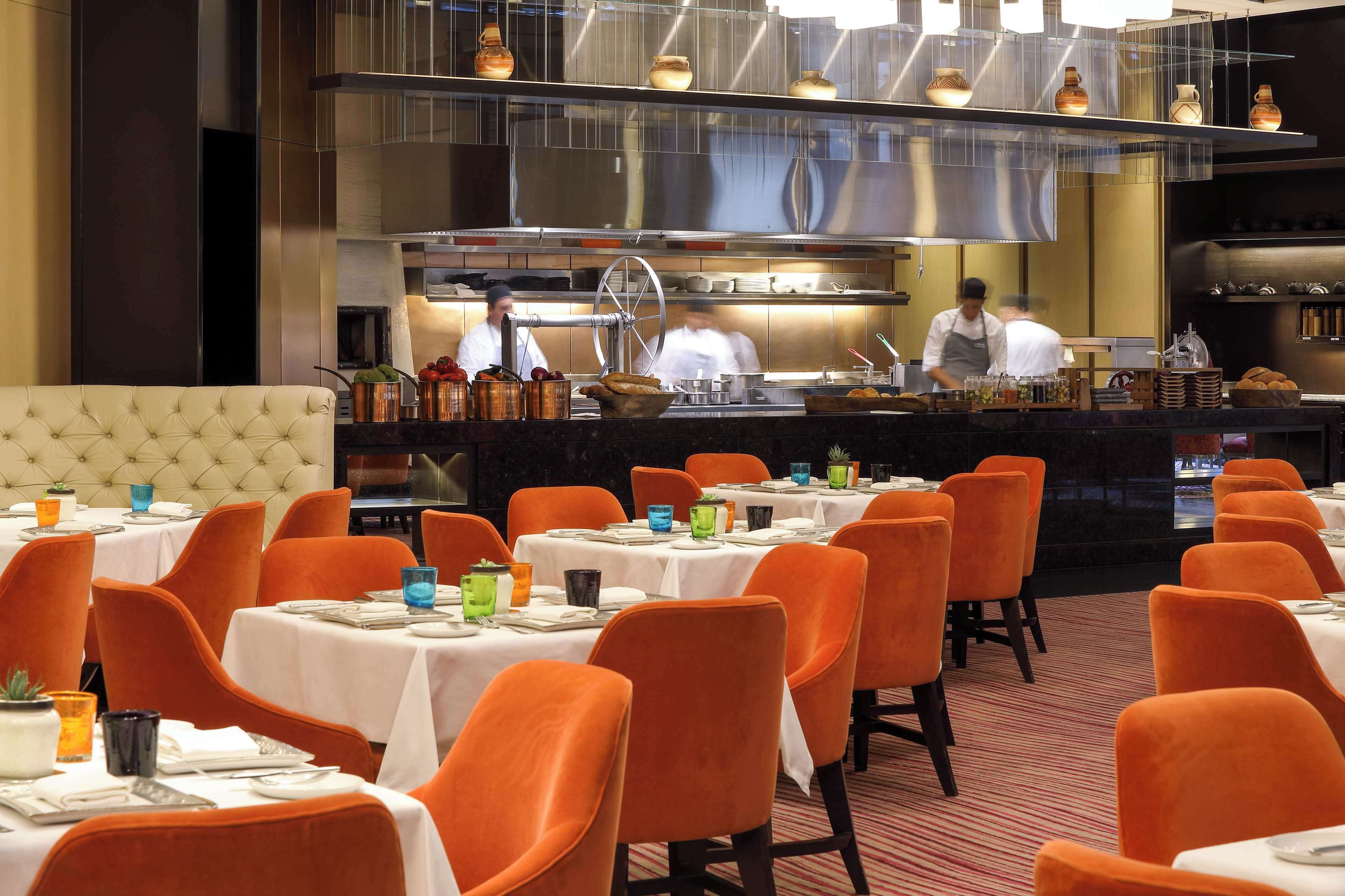 A restaurant with tables set for dining, orange chairs, and a kitchen area with chefs working.