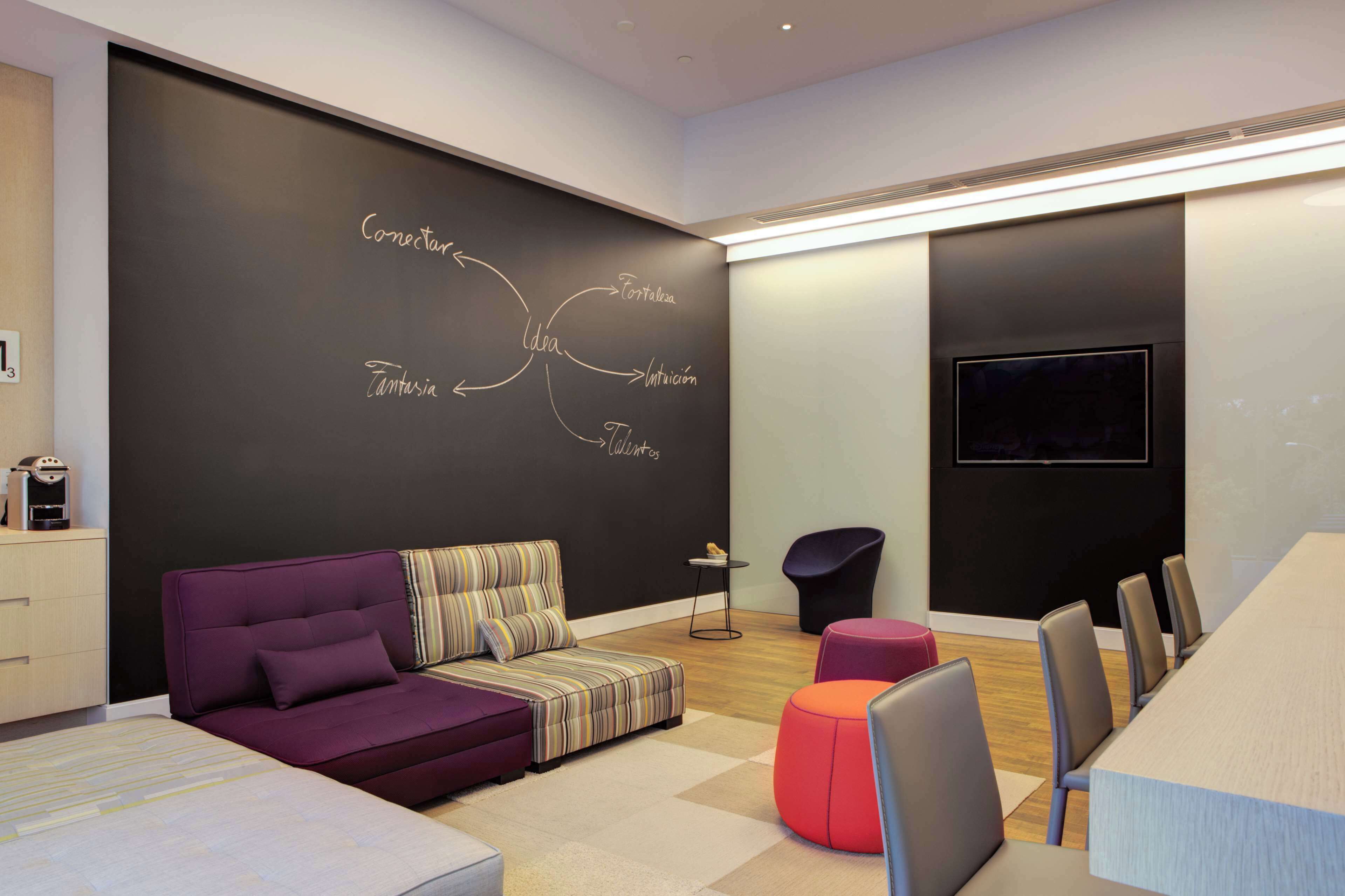 Ein moderner Besprechungsraum mit einer Tafel, auf der Wörter wie „Idee“, „Innovation“ und „Kreativität“ stehen. Es gibt ein violettes Sofa, einen kleinen Tisch und einen Konferenztisch mit Stühlen.