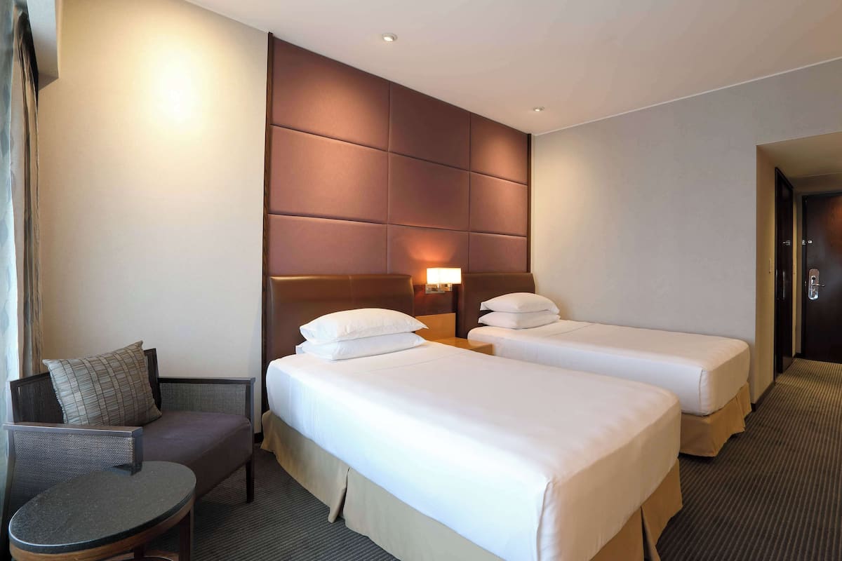 Une chambre d’hôtel avec deux lits simples, un canapé, une petite table ronde et une tête de lit dotée d’un luminaire.